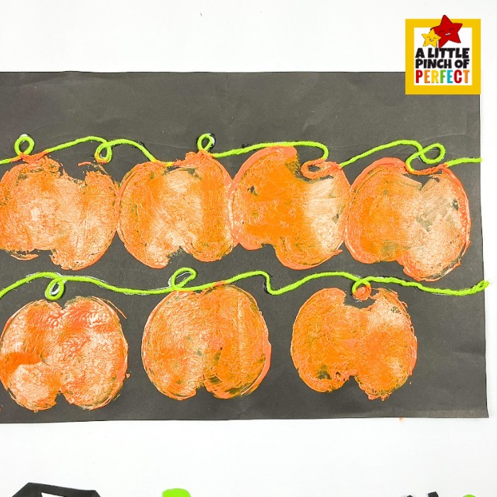 Apple stamped pumpkin patch Halloween art project for kids — the perfect Halloween process art activity for preschool, kindergarten, or homeschool fall unit studies! Orange apple-stamped pumpkin prints are arranged in two cheerful rows on black paper, with green yarn glued along the wavy vine lines to connect them into a charming pumpkin patch.